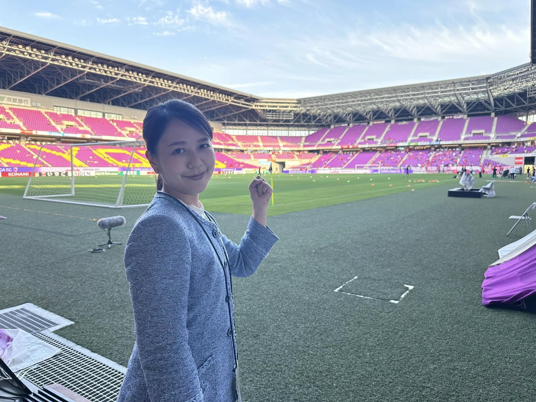 A woman in a grey suit smiles at the camera and holds her fist up in a soccer stadium