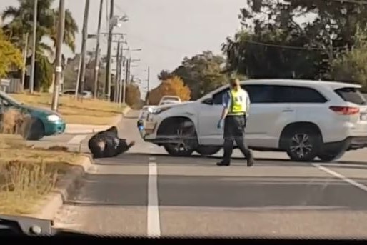 A white SUV is parked on a suburban street with a man falling to the ground in front of it.