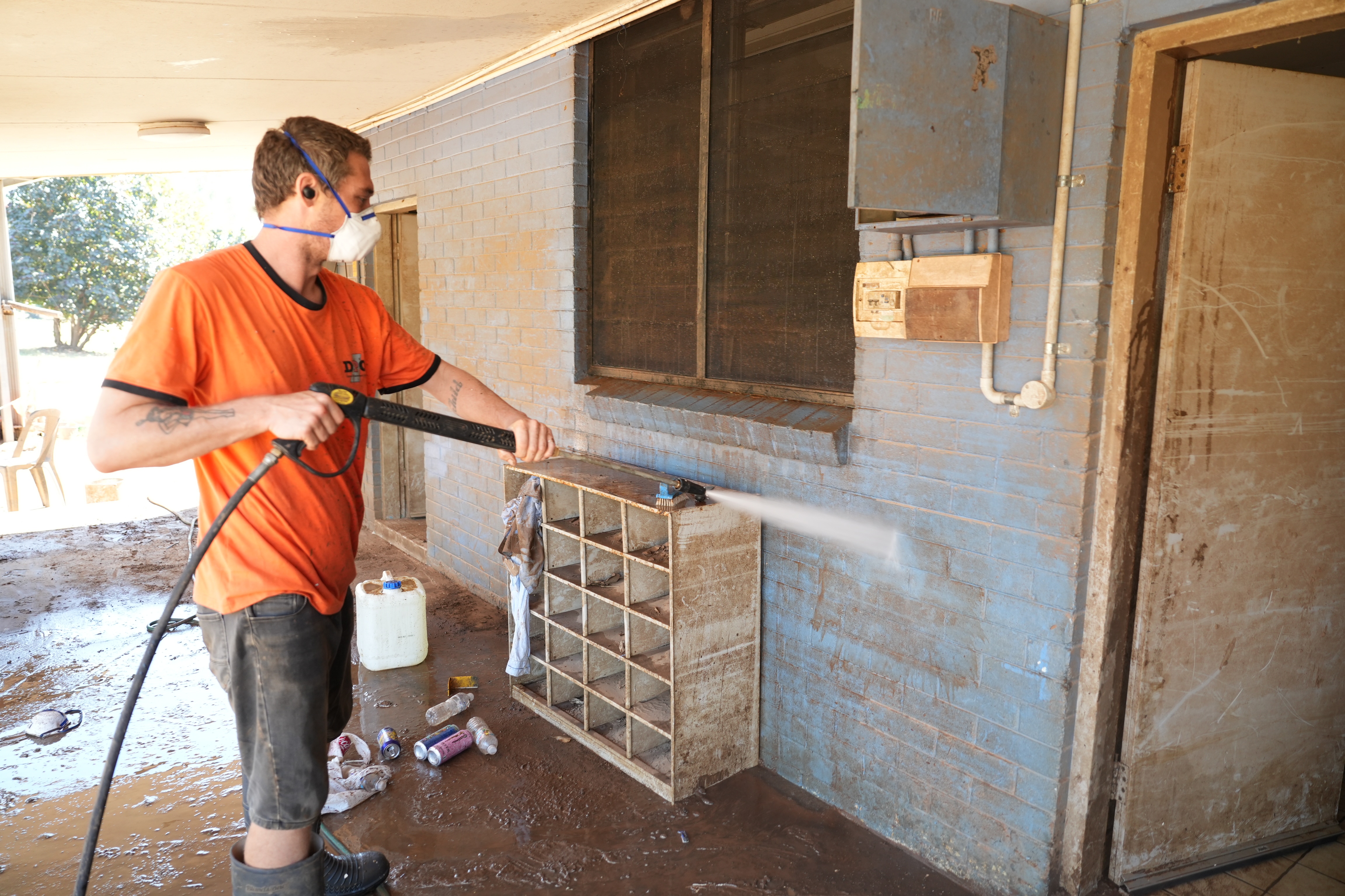 A man in a bright orange shirt sprays the muddy blue wall of a house