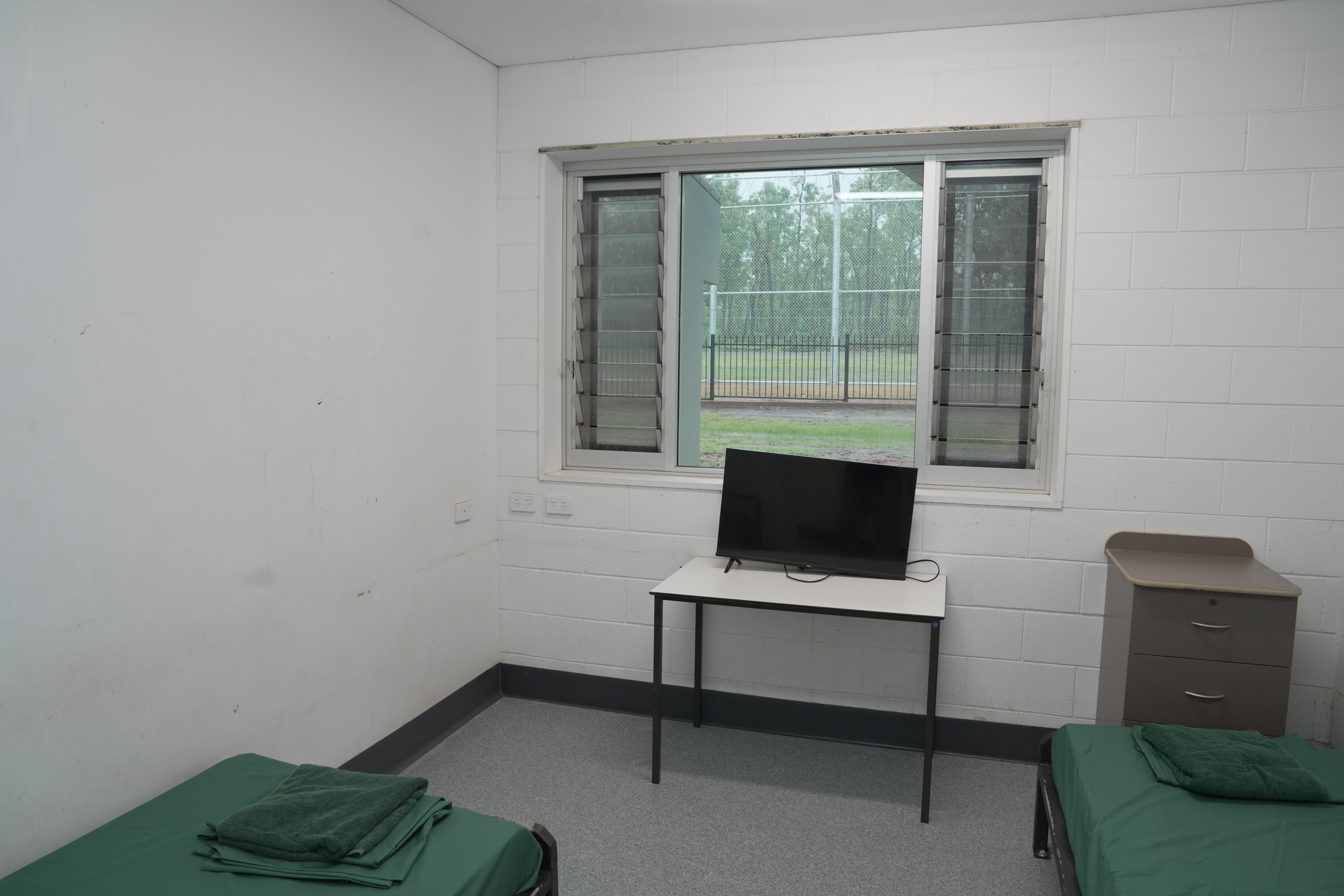 Dos camas individuales en los bordes del marco, sábanas verdes. Televisor sobre mesa blanca, con una pata quitada. Estantes al final de una cama.
