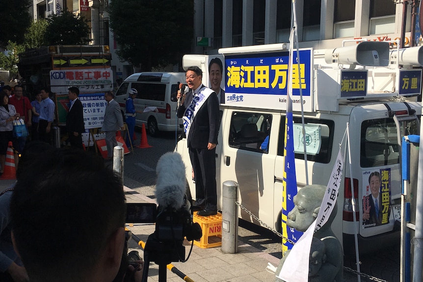 A candidate gives a speech while standing on an orange box in front of a white van parked on the street.