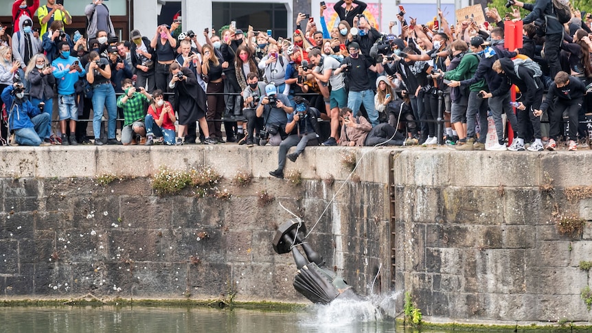 A crowd of people stand on a canal's edge as a statue falls into the water below 