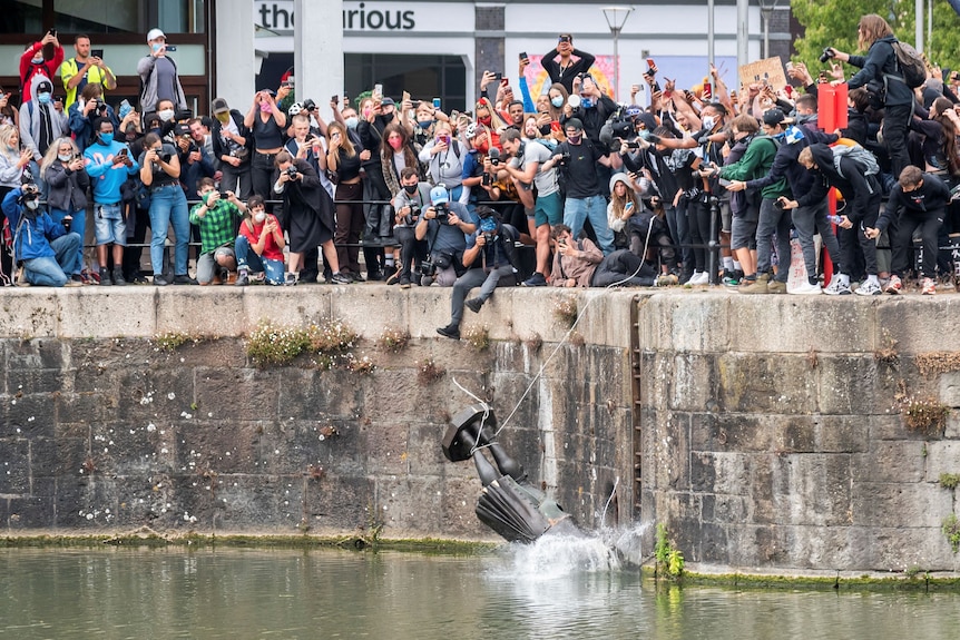 A crowd of people stand on a canal's edge as a statue falls into the water below 