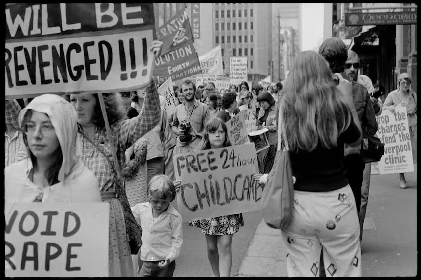 Black and white picture of a protest focused on a kid with a placard reading "free 24 hour childcare"