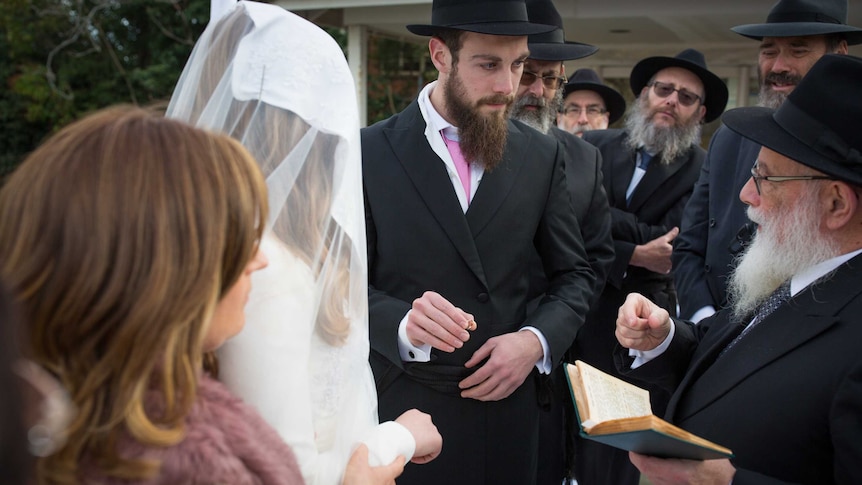 A groom surrounded by older Jewish men holds a ring towards the hand of his bride, head covered.