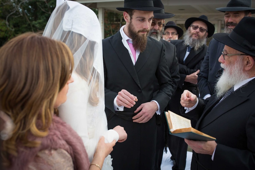 A groom surrounded by older Jewish men holds a ring towards the hand of his bride, head covered.