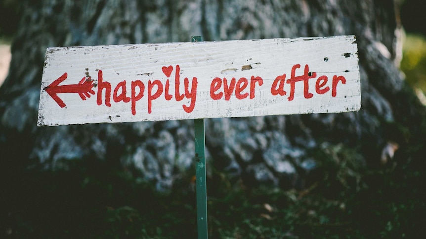 A hand-painted sign saying "happily ever after" is stuck into the ground in front of a tree.