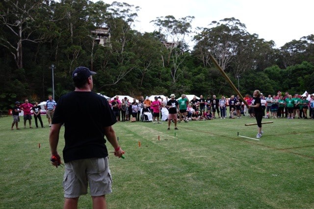 A woman launches a tall wooden pole at a park among a row of spectators