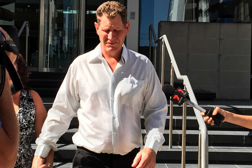 A mid-shot of a man wearing a white button-up shirt walking away from Perth District Court with reporters and cameramen nearby.