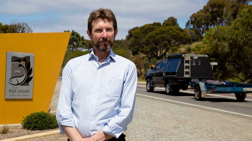 A man with short brown hair stands on the side of a highway as a car with a trailer drives past.