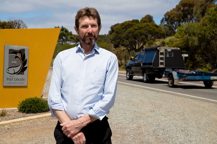 A man with short brown hair stands on the side of a highway as a car with a trailer drives past.