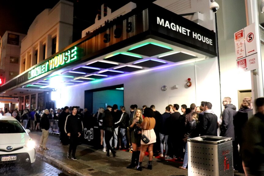 Une file d'attente de personnes devant une boîte de nuit avec une enseigne au néon disant 'Magnet House'.