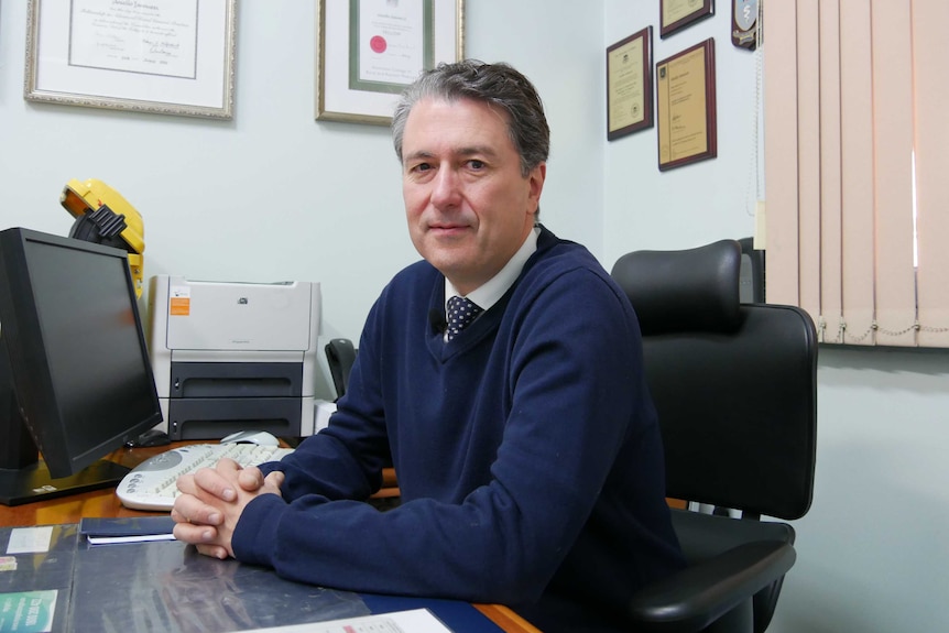 A middle-aged man in a blue sweater sits at a desk with has hands clasped.