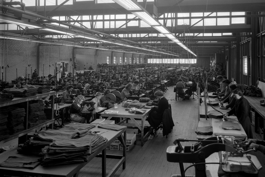 A black and white image of hundreds of people in a factory working on tables. 