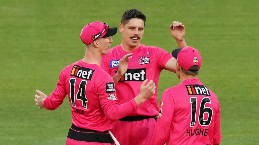Ben Dwarshuis holds his left arm up while two other people wearing pink cricket clothes stand in front of him