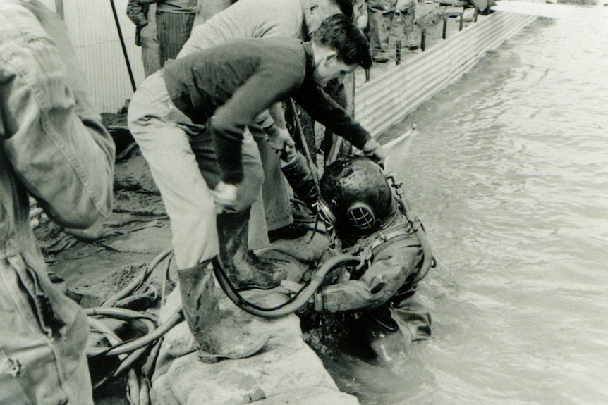 A man wearing a diving suit and going under water. There is a man on the surface helping him out.