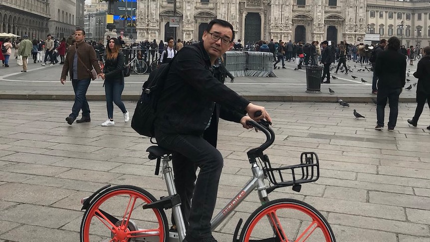 Yang Hengjun sits on an orange-wheeled bike wearing all black with a black backpack on a grey day with a church in background.