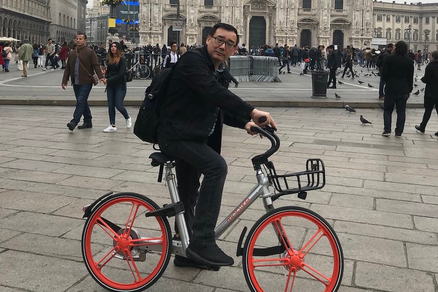 Yang Hengjun sits on an orange-wheeled bike wearing all black with a black backpack on a grey day with a church in background.