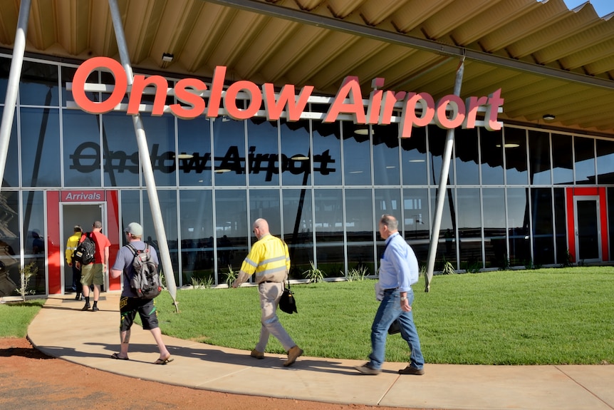 A group of men carry bags towards the entrance for an airport terminal. The sign reads "Onslow Airport"