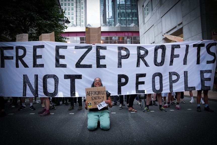 A woman kneels in front of a banner reading "Freeze profits, not People" 