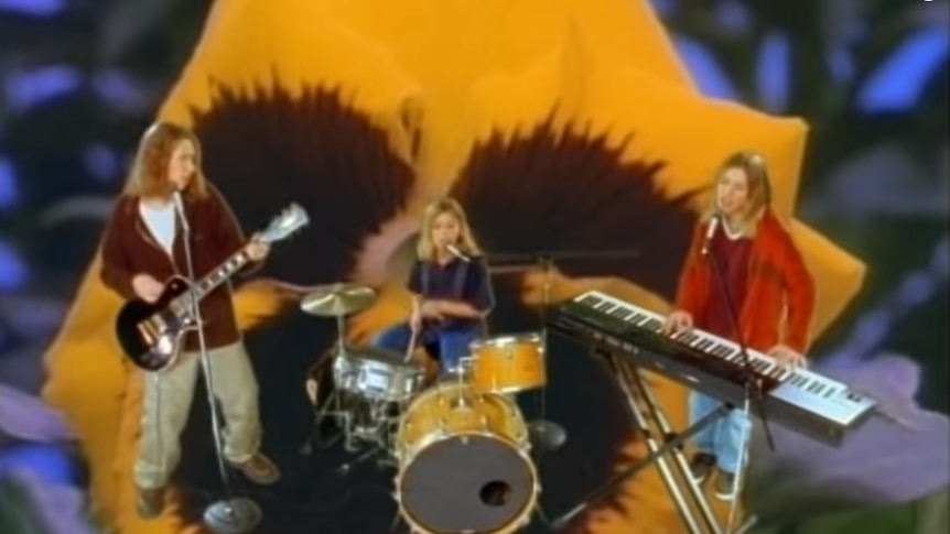 Three boys play guitar, drums and keyboard in front of a backdrop of a giant flower