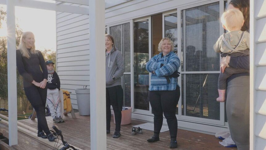 A group of ladies in wetsuits and children, on a porch before surf