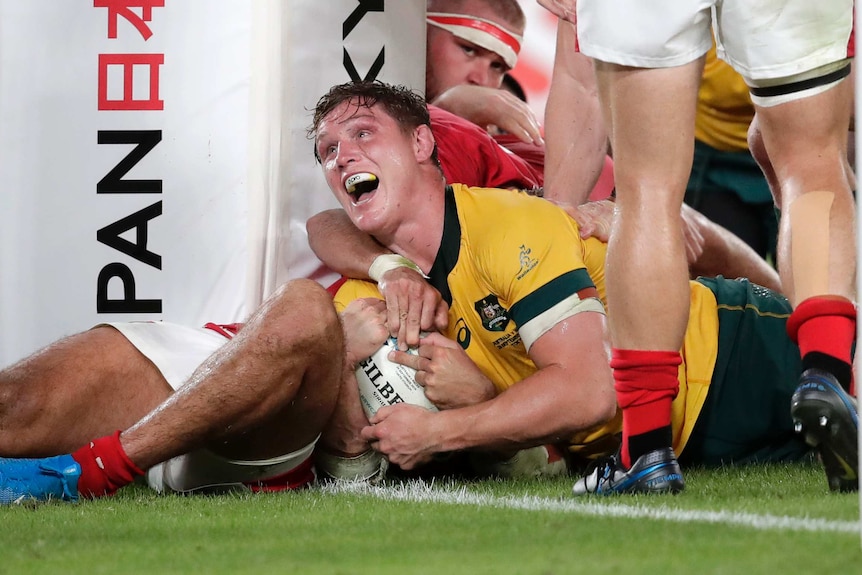 A male rugby union player looks up with a smile on his face after scoring a try under the goal posts.