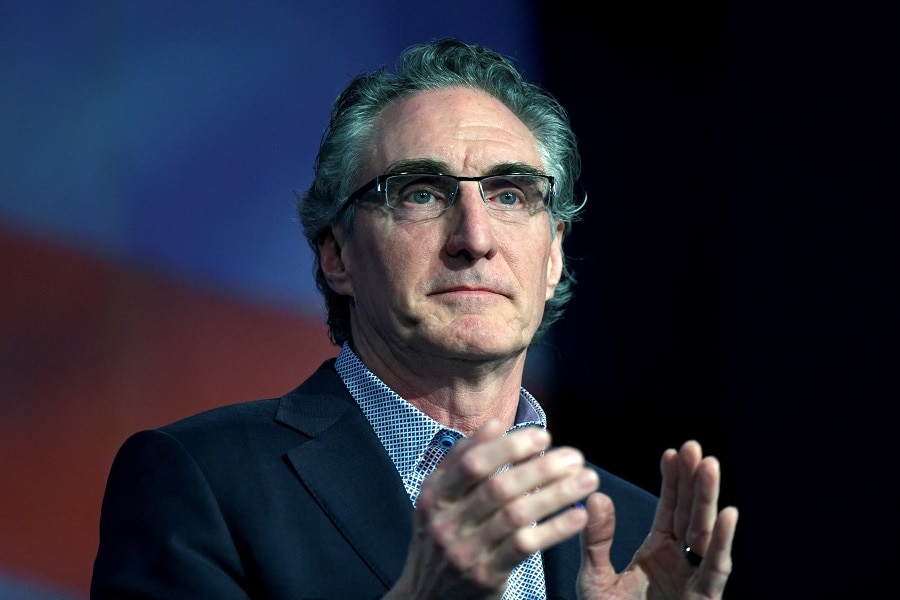 An older white man with glasses and an angular face claps on stage in front of a red and white backdrop.