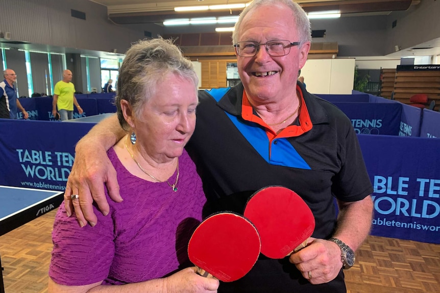 An elderly couple hug while holding table tennis paddles