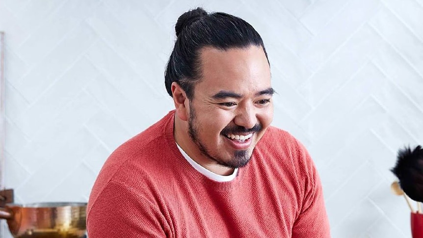 Adam Liaw wears a pink jumper, standing in front of a white wall, smiling off camera