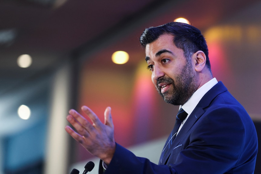 A man in a blue suit gestures as he speaks at a lectern