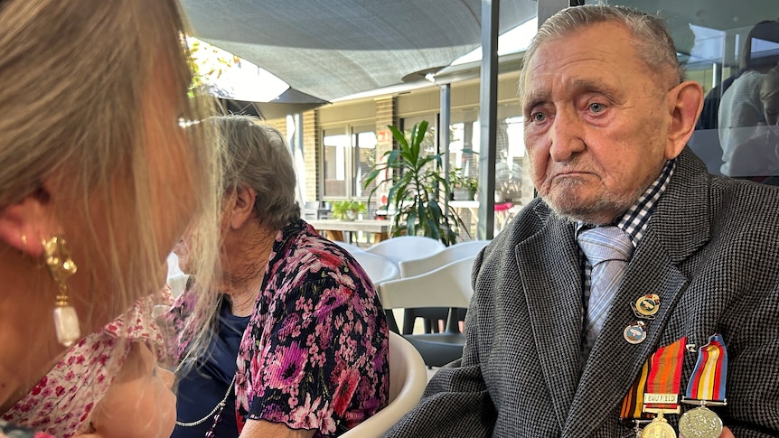 An elderly man sits with a sad expression on his face wearing a coat displaying war medals