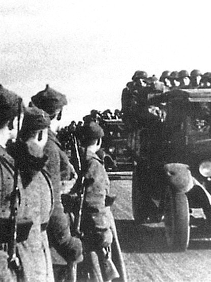 Soldiers stand to attention as cars full of soldiers drive by.