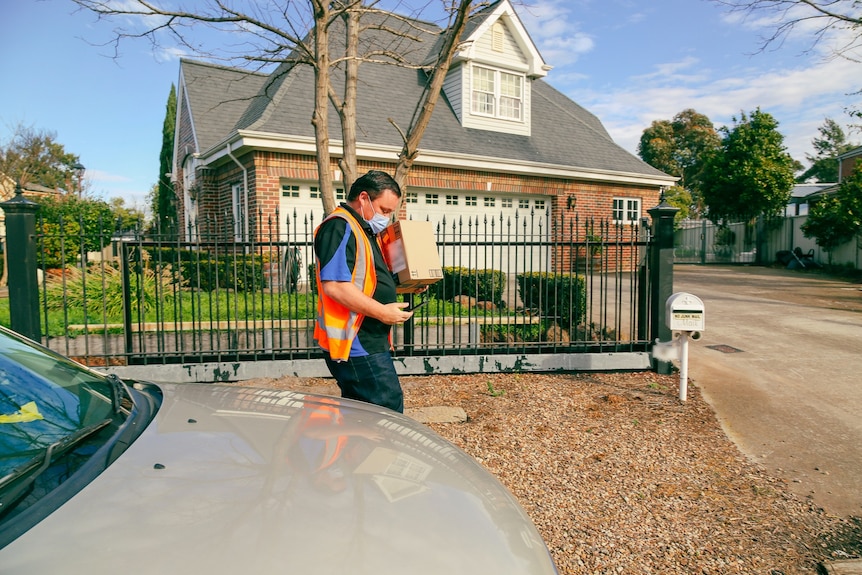 A man in an orange high-vis vest carries a box as he walks past a house on a suburban street. 