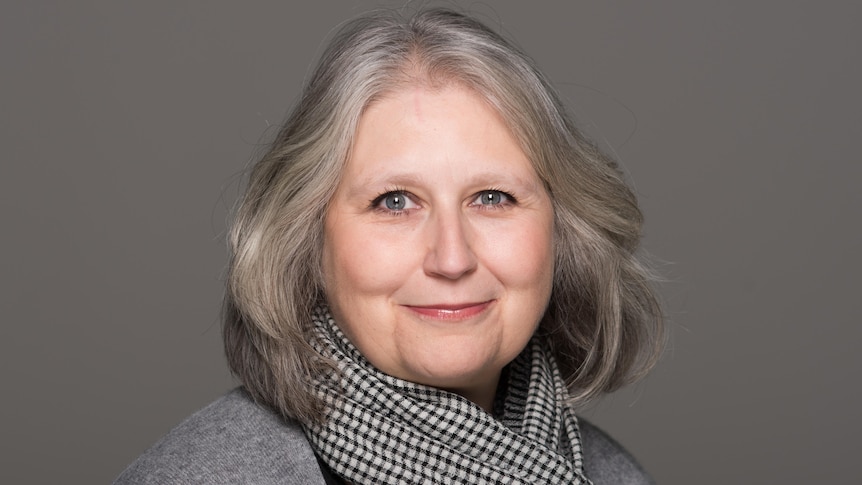 A headshot photo of a white middle-aged woman with short grey hair wearing a checkered scarf and grey cardigan.