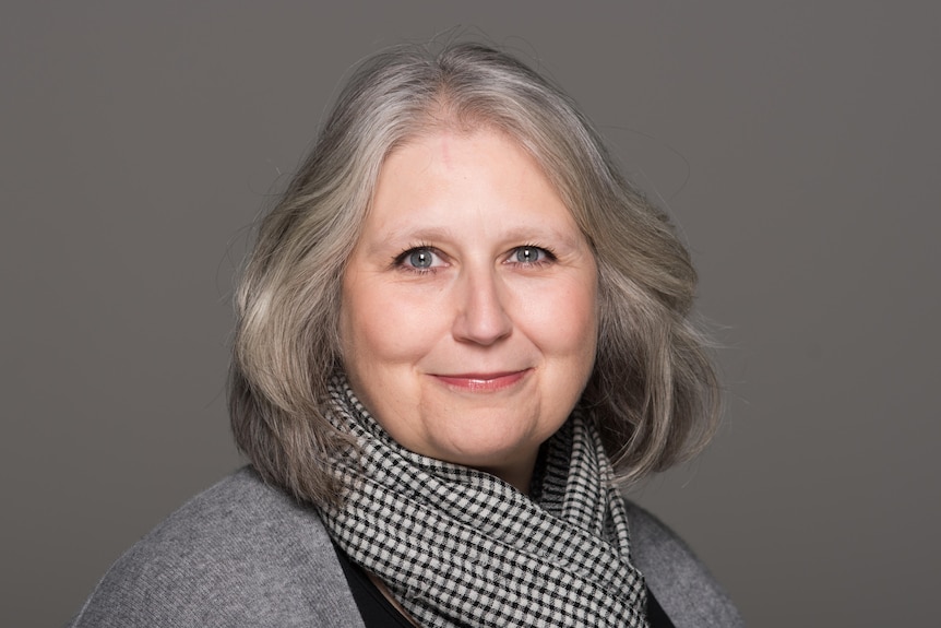 A headshot photo of a white middle-aged woman with short grey hair wearing a checkered scarf and grey cardigan.