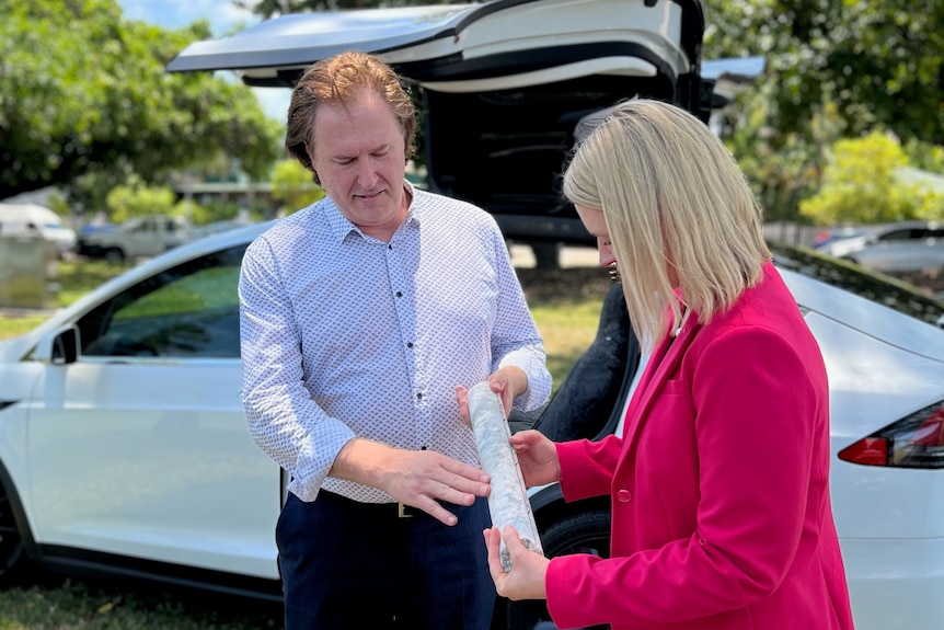 a man showing a mining sample to a woman with an electric car in the background.