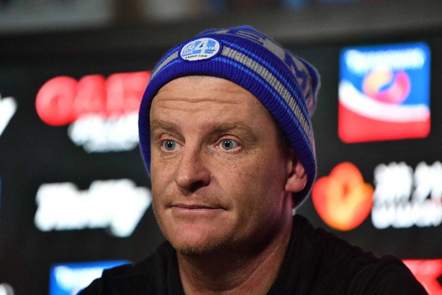 Man in a beanie sits at a table in a press conference.