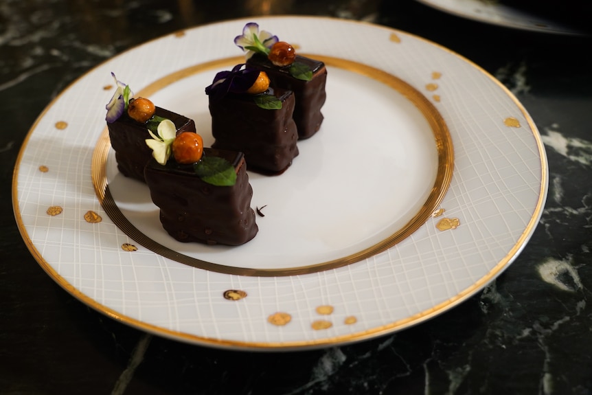 Four small chocolate-covered desserts sit on a white plate with gold accents
