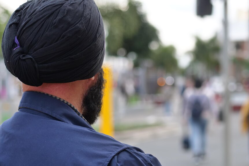 A man wearing a black turban and blue shirt looking out over a city street crossing 