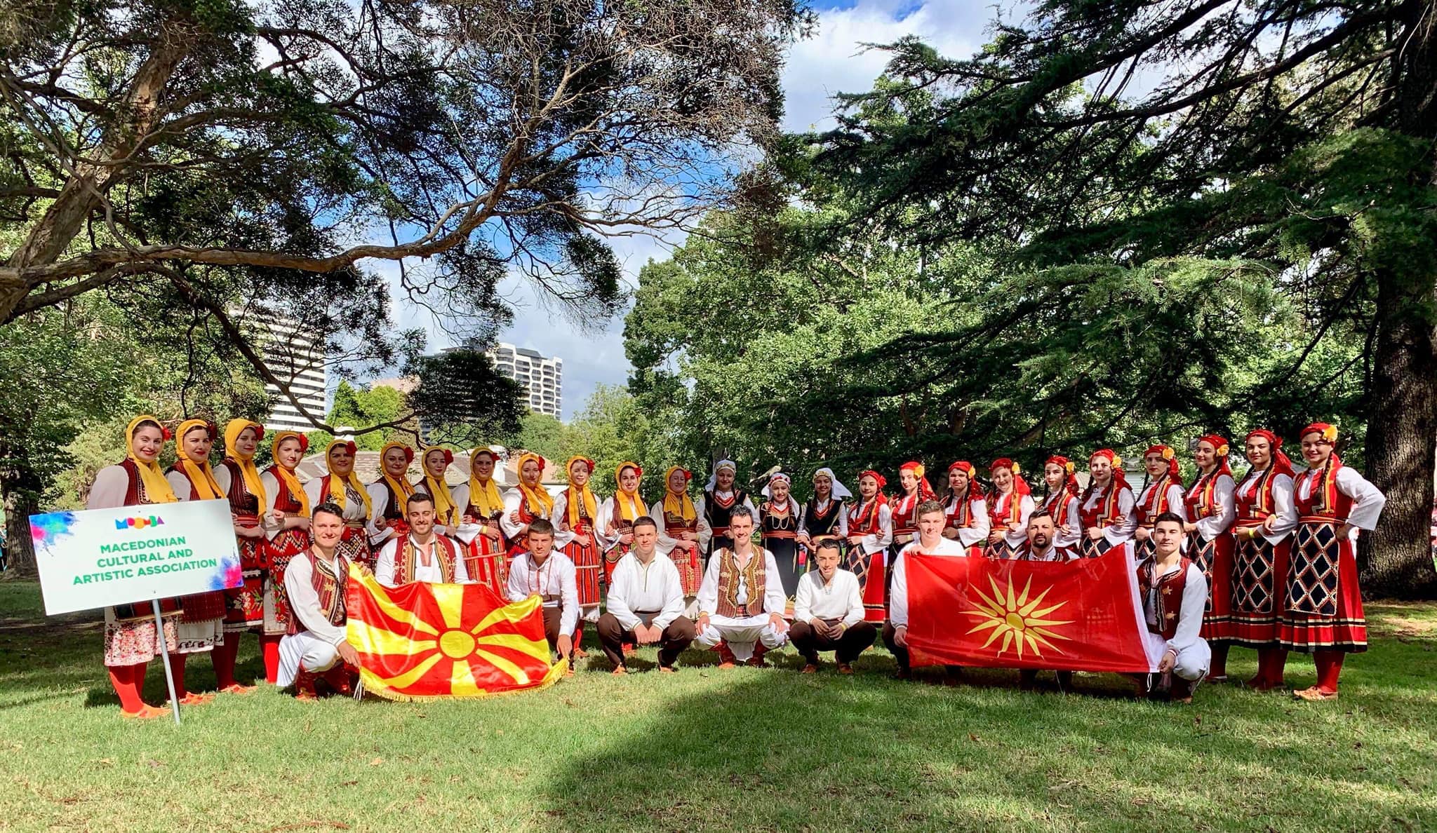 Un grupo de hombres y mujeres con ropa tradicional macedonia posan para una fotografía con dos banderas.