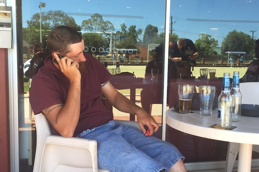 A man on a mobile phone sits at a bowling club with beer beside him