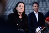 An AFL football executive stands at a lectern as she answers questions, while a colleague stands behind and off to the side.  