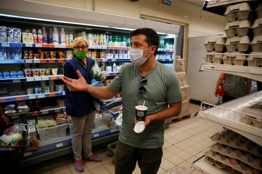 A man in a green t shirt wears a face masks as he gestures with his right palm up and holds a drink in his left hand.