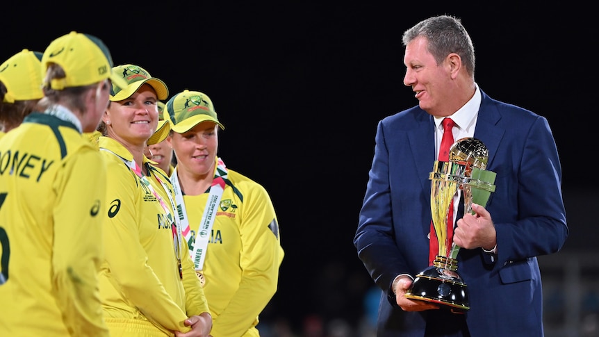 A man in a suit presents a trophy to women's cricket players wearing yellow and green