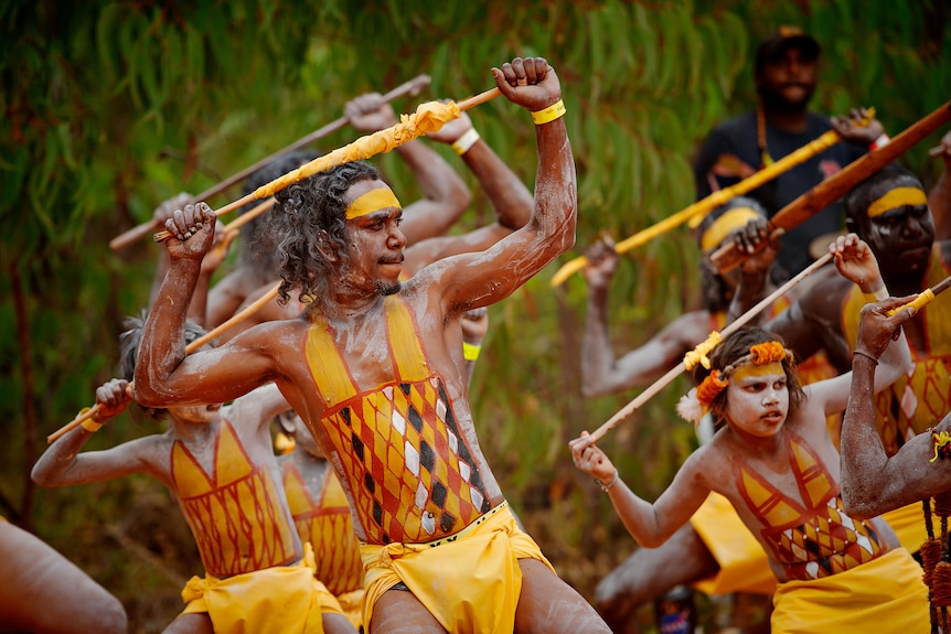 Аборигенски танцьори, боядисани в жълто, черно, червено и бяло, изпълняват танц, държейки инструмент над главите си