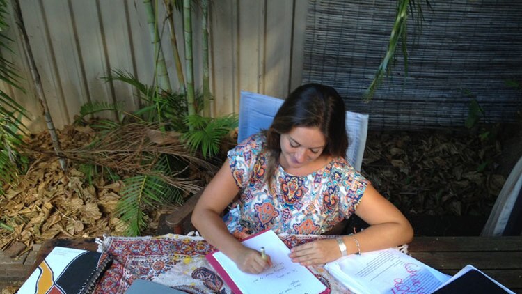 A woman writes notes beside a laptop in a tropical garden setting