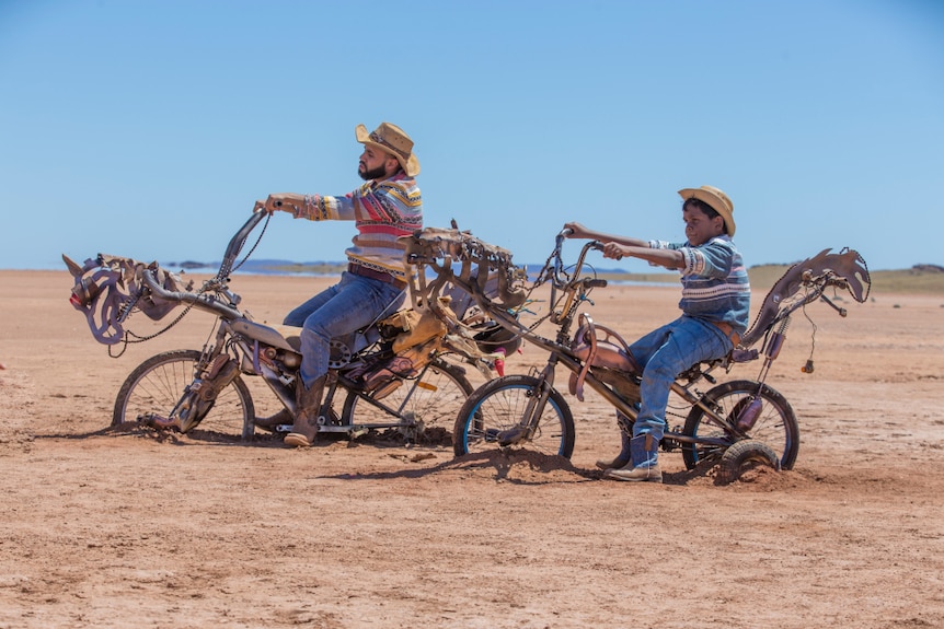 A man and boy wear farmer's hat, blue jeans, boots and shirt and sit on modified bicycles in the desert on a sunny blue sky day.