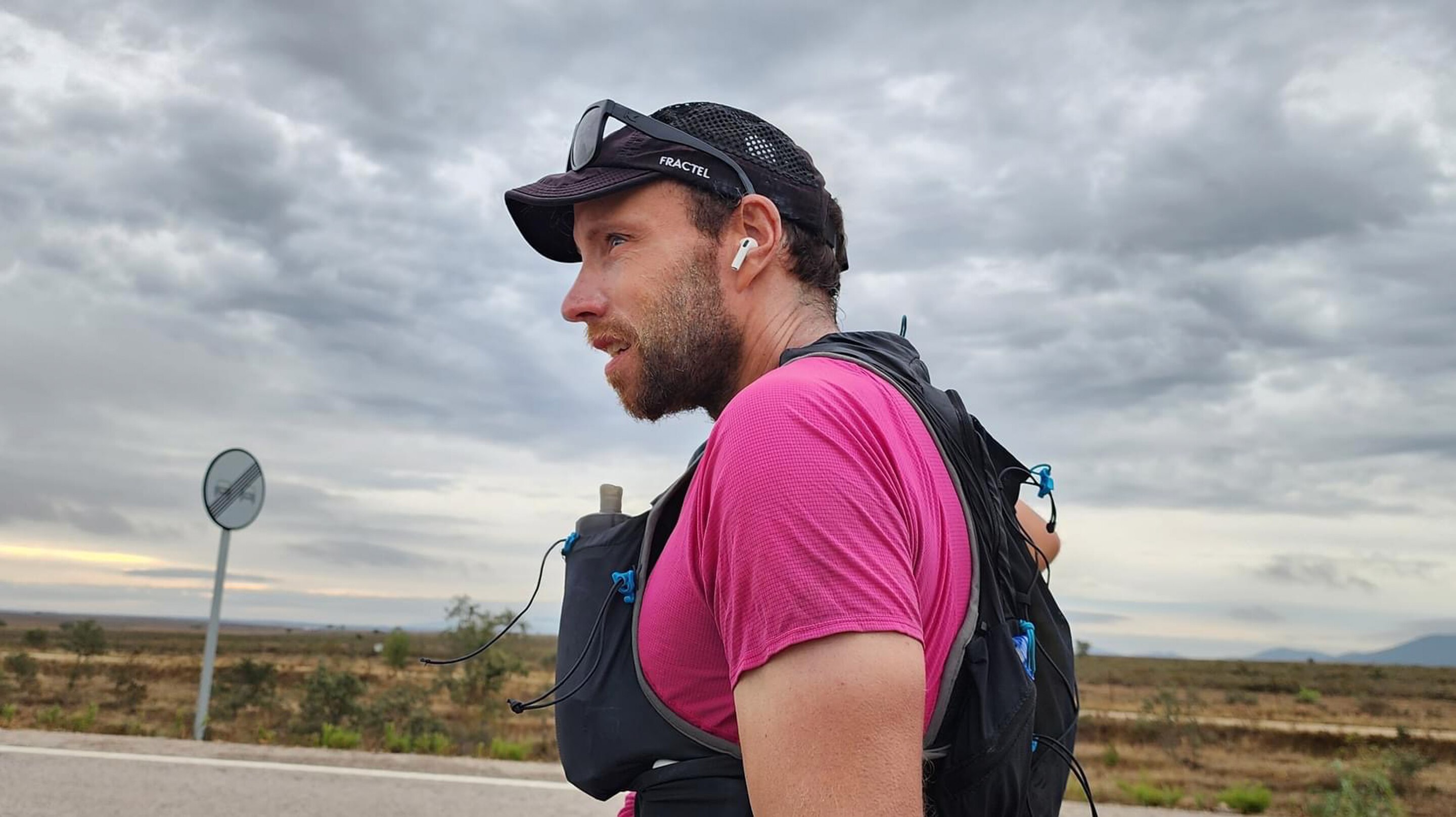 Tight side profile of a man in a black cap, red top and black running vest walking down the road with a determined expression.