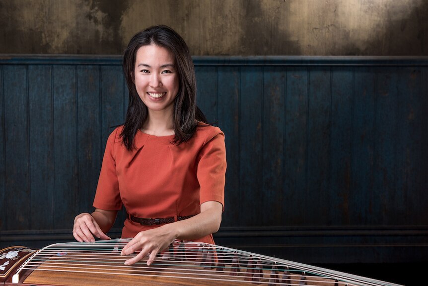 Zhao Liang plays a guzheng and smiles at the camera. She wears a red dress.